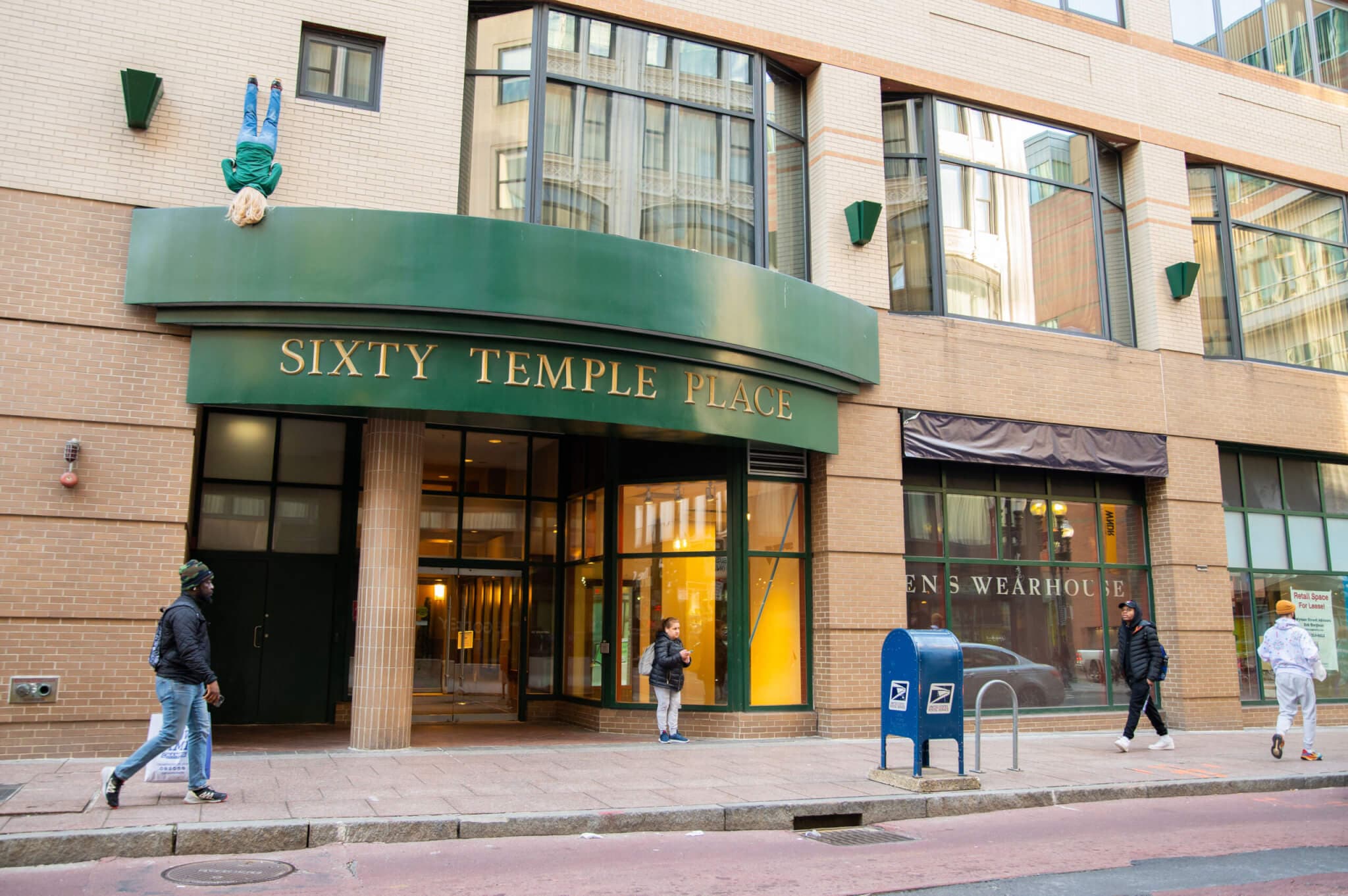 People walk past a building in Downtown Crossing with a sculpture of an upside down man hanging off the top.